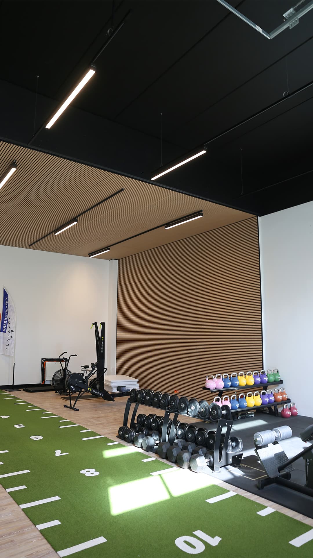 Trainingshalle 2 im September: Holzlamellendecke und -wand mit Lichtleisten unter schwarzer Decke, grüne Sprintbahn mit Metermarkierung, Kurzhantelablage, farbige Kettlebells, Rudergerät und Skiergometer an der Rückwand.